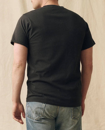 A person wearing THE GREAT. FALL 23 MEN The Men's Pure Knits Slim Tee in Solid -- Almost Black and blue jeans stands facing away from the camera against a beige fabric backdrop.