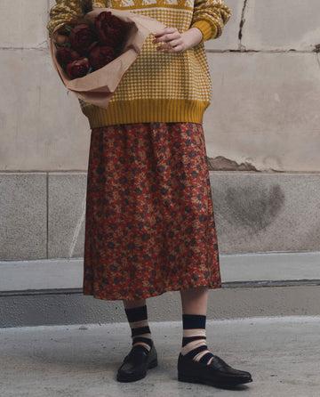 Wearing THE GREAT.'s The Canary Skirt from Gilded Poppy, a person stands against a weathered concrete wall, their lower half visible beneath a patterned yellow sweater and a bouquet of dark red flowers in hand.