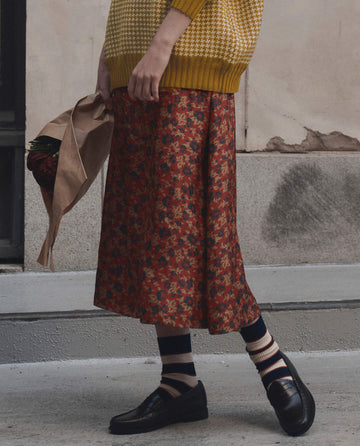 A person wearing a yellow sweater, THE GREAT. F26 D1 The Canary Skirt by Gilded Poppy, striped socks, and black loafers stands on the pavement, holding a brown paper bag filled with strawberries.