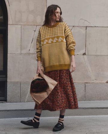 A person walks along a city sidewalk holding a bouquet wrapped in brown paper, dressed in a yellow patterned sweater, black shoes, and The Canary Skirt from THE GREAT. F26 D1 – Gilded Poppy, featuring red floral accents.