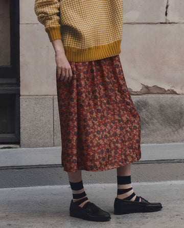 A person is wearing THE GREAT. F26 D1 The Canary Skirt from Gilded Poppy, styled with a yellow patterned sweater, striped socks, and black loafers as they stand on a sidewalk next to a stone building.