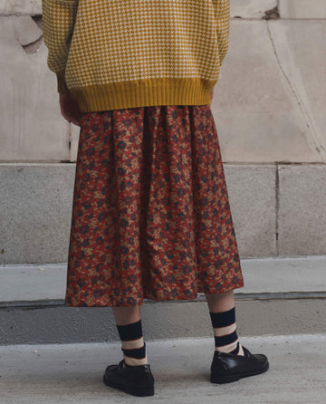 A person stands facing a textured wall, wearing THE GREAT. F26 D1’s The Canary Skirt by Gilded Poppy, paired with a yellow houndstooth sweater, striped socks, and black shoes.