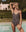 A woman stands poolside outdoors in THE GREAT. SP25 SWIM The Reversible Lace Up One Piece -- Leopard and Black, with one arm raised above her head. Lush greenery and a light-colored building are visible in the background.