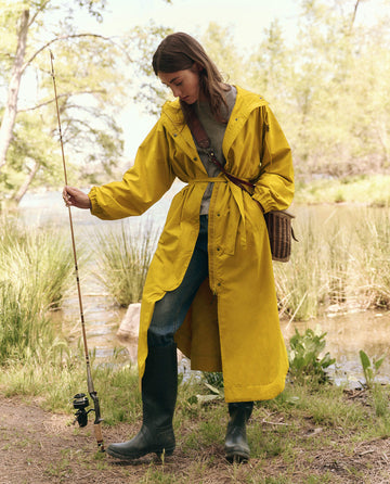 Wearing The Rain Trench by THE GREAT. SP25 TGO in Sunshine and matching boots, a person holds a fishing rod lakeside amid lush trees and grass.