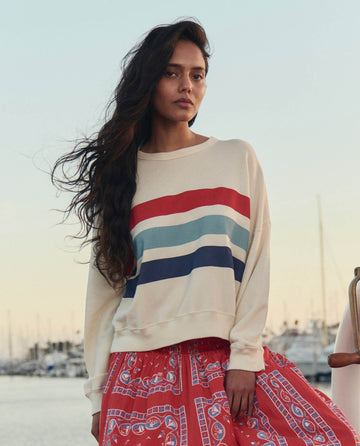 A woman with long hair wears THE GREAT. SU26 The Teammate Sweatshirt W/ Saltwater Stripe in Washed White and a red patterned skirt, standing outdoors by a marina at sunset.