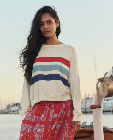 A woman stands on a dock by boats, wearing THE GREAT. SU26’s The Teammate Sweatshirt W/ Saltwater Stripe in Washed White and a red patterned skirt.