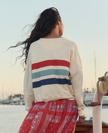 Wearing THE GREAT. SU26's The Teammate Sweatshirt W/ Saltwater Stripe in Washed White, a person with long hair stands near water and boats, paired with a red patterned skirt and facing away.