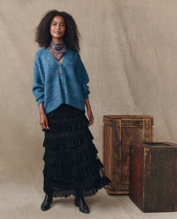 Indoors, a woman wears THE GREAT. H25 COLLECTION’s The Brushed Melange Cardigan in Wintertide Blue, styled with a patterned scarf, black tiered skirt, and black boots, beside two wooden crates against a neutral backdrop.