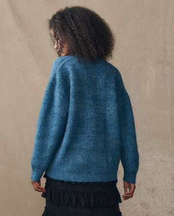 A person with curly hair stands with their back to the camera, wearing THE GREAT. H25 COLLECTION’s The Brushed Melange Cardigan in Wintertide Blue and a black ruffled skirt, set against a plain beige background.