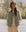 A person stands by a lake wearing THE GREAT. SP25 TGO's The Reversible Ultra Light Down Vest in Falling Daisy and Cream over a light shirt, with trees and mountains in the background.