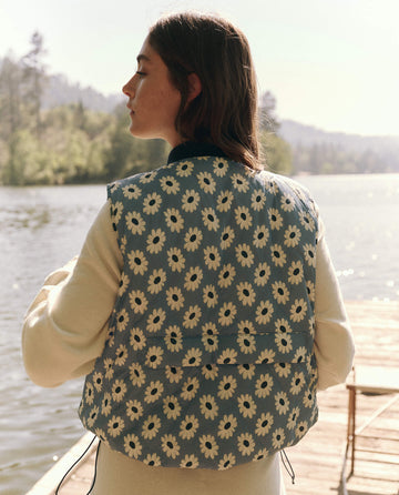 On a wooden dock by the lake, a person wears The Reversible Ultra Light Down Vest in Falling Daisy and Cream from THE GREAT. SP25 TGO over a floral long-sleeve shirt, with trees and hills in the background.