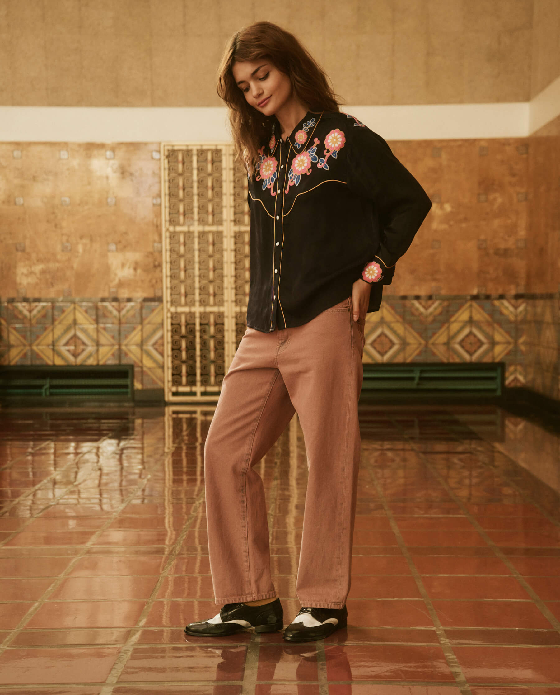 Person standing in a tiled room wearing a black shirt with floral embroidery, "The Billy Jean. Clean Hem -- Musky Pink" pants from THE GREAT. PS24 SALE, and black and white shoes. The room has patterned walls and a decorative radiator cover, creating an atmosphere reminiscent of relaxed silhouettes from vintage-inspired denim days.