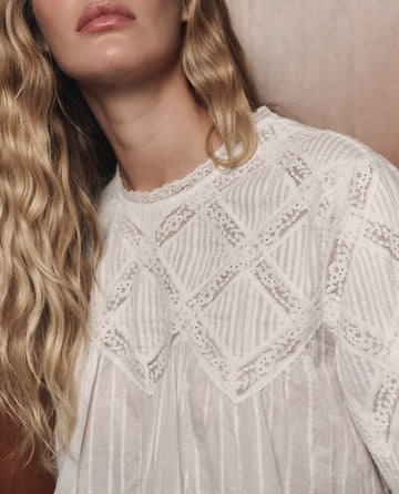 A woman with wavy blonde hair wears The Lattice Lace Victorian Top in white by THE GREAT. F26 COLLECTORS, featuring a sheer geometric lace pattern, standing against a brown background.