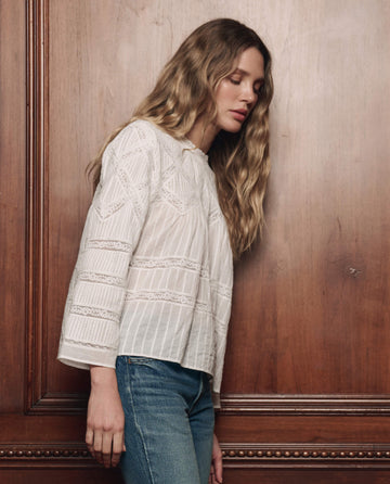 A person with long, wavy hair wearing THE GREAT. F26 COLLECTORS' The Lattice Lace Victorian Top in white and blue jeans stands against a wooden wall, looking down.