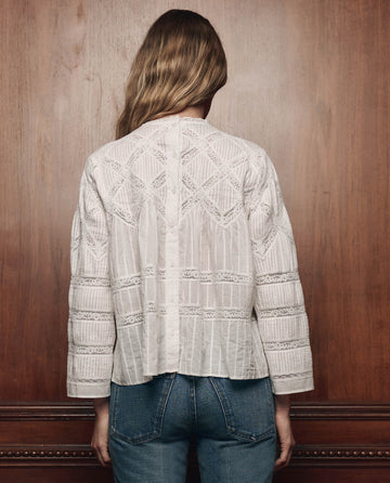 A person with long hair stands facing a wooden wall, wearing THE GREAT. F26 COLLECTORS’ The Lattice Lace Victorian Top in white, featuring intricate patterns and blue jeans.