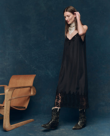 A woman in THE GREAT. SP25 COLLECTORS The Legacy Dress—Black, cowboy boots, and a white neck scarf stands by a wooden chair against a dark blue backdrop, creating a timeless fall look.
