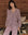 A person wearing The Pajama Shirt in Bordeaux Plaid from THE GREAT. HOL 24 SLEEP stands by a bed with a wooden background, showcasing cozy sleepwear.