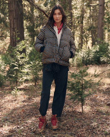 A person stands in a forest wearing THE GREAT. F26 TGO The Polar Puffer. -- Black Paisley, matching pants, red hiking boots, and a red bandana around their neck.