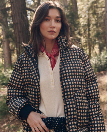 A woman stands in a forest wearing The Polar Puffer. -- Black Paisley by THE GREAT. F26 TGO, a light henley shirt, and a red neckerchief, looking at the camera.