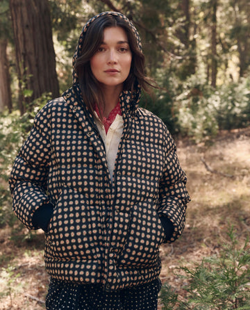 A woman wearing The Polar Puffer. -- Black Paisley by THE GREAT. F26 TGO stands outdoors in a wooded area, looking at the camera.