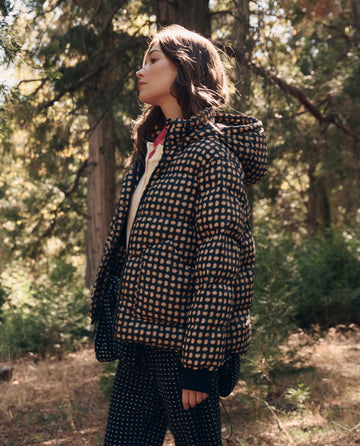 A person wearing The Polar Puffer. -- Black Paisley by THE GREAT. F26 TGO stands outdoors in a sunlit forest, looking to the side.
