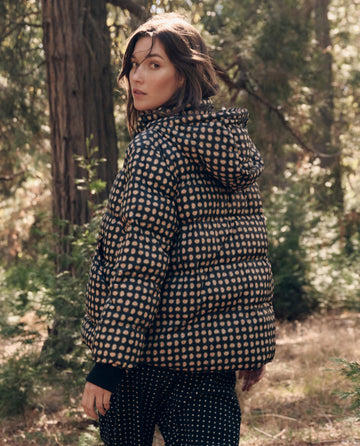 A person in THE GREAT. F26 TGO The Polar Puffer. -- Black Paisley with polka dot pants stands in a forest, glancing back over their shoulder.