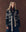 A woman stands against a brown background, wearing THE GREAT. F25 D1 The Reversible Shepherd Cardigan in navy and cream with geometric patterns and a matching belt, layered over a long dark skirt and patterned scarf.