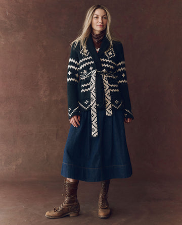 A woman stands against a brown backdrop wearing THE GREAT. F25 D1 The Reversible Shepherd Cardigan in Navy And Cream, styled with a long denim skirt, brown lace-up boots, and a scarf around her neck.