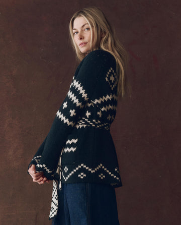 A person with long blonde hair models THE GREAT. F25 D1 The Reversible Shepherd Cardigan in navy and cream over blue jeans, posing against a brown textured background.