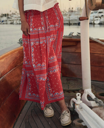 A person stands on a wooden boat wearing THE GREAT. SU26 The Canary Skirt in Red Sailboat Bandana, paired with beige boat shoes, with a marina and boats in the background.