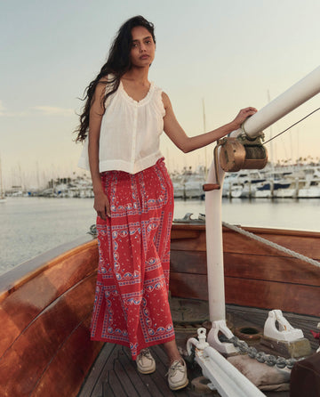 A woman stands on a wooden boat in front of a marina, wearing THE GREAT. SU26’s The Canary Skirt and the Red Sailboat Bandana for a vibrant, stylish look.