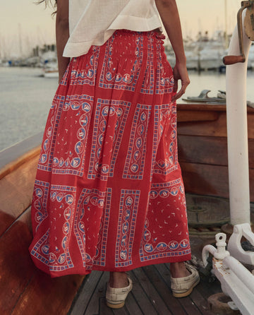 A person in a white top and THE GREAT. SU26 Canary Skirt with a Red Sailboat Bandana stands on a wooden boat, marina and docked boats in the sunset background.