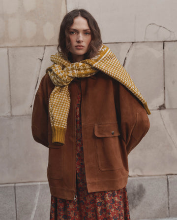 A woman wearing THE GREAT. F26 D1 The Suede Folsom Jacket in Cognac, paired with a red floral dress and a yellow patterned scarf, stands against a textured, cracked wall.