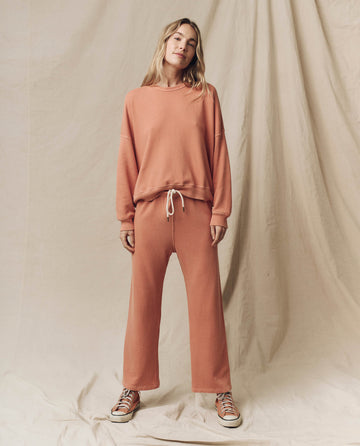 A person stands against a fabric backdrop wearing THE GREAT. SU24 KNITS' oversized, Sun Dried Orange relaxed fit Teammate Sweatshirt with vintage details and matching wide-legged pants with drawstrings, paired with sneakers.