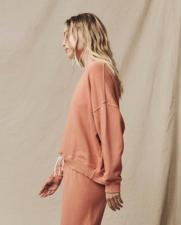 A person with long, wavy hair is wearing The Teammate Sweatshirt in Sun Dried Orange by THE GREAT. SU24 KNITS, paired with matching pants that feature vintage details. They are facing left against a plain, beige background.