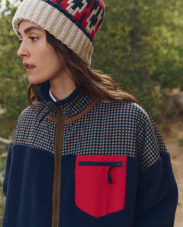 A woman stands before greenery, wearing THE GREAT. F25 TGO The Print-Mix Plush Terrain Full-Zip in Mini Frosted Daisy & Navy, featuring a patterned knit hat and cozy welt pockets with a red zippered chest pocket.
