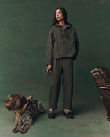 A woman wears THE GREAT. F25 D2's The Utility Pullover in Dusty Moss, standing indoors with a shaggy brown dog on a leash by her side, next to a brown leather chair and set against a rich green backdrop.