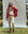 Dressed in a casual ensemble, a person wears a red jacket over The Varsity College Sweatshirt by THE GREAT. PS25 in washed white with crest graphic and joggers, standing on grass before a light blue backdrop with trees and bushes visible behind them.