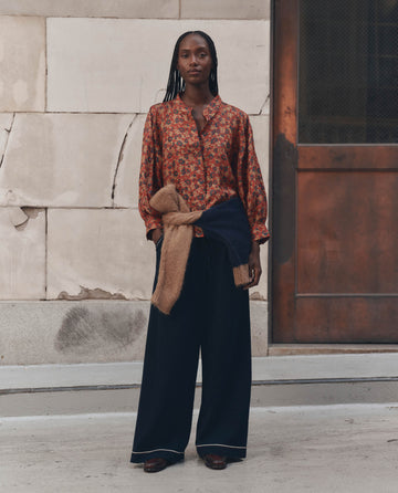 A woman stands outdoors in the Ashland Top — Gilded Poppy by THE GREAT., styled with wide-leg dark pants and two sweaters tied at her waist, against a stone wall and metal door backdrop.