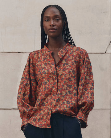 A person with long braided hair wears THE GREAT. F26 D1 The Ashland Top in Gilded Poppy—an airy, red floral-patterned women’s top—styled with dark pants and standing against a light, textured wall.