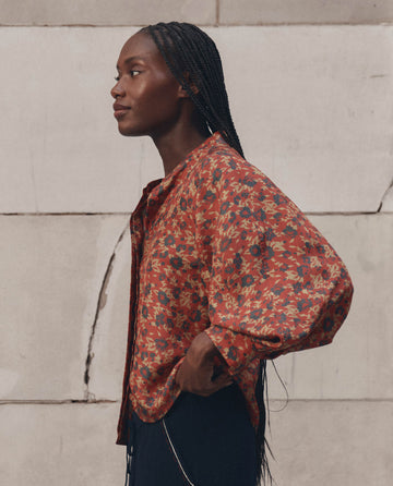 A woman with braided hair stands in profile, wearing THE GREAT.’s Ashland Top in Gilded Poppy—a long-sleeved, red floral-patterned blouse—set against a textured, light-colored stone wall.