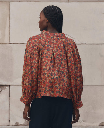 A person with braided hair stands facing away from the camera, wearing THE GREAT.'s Ashland Top in Gilded Poppy—a rust-colored, floral women's top with gathered cuffs—paired with dark trousers against a stone block wall.