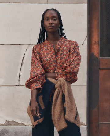 A woman with long braids stands in front of a worn wall, wearing THE GREAT.'s The Ashland Top in Gilded Poppy, paired with dark pants and a tan sweater tied around her waist.