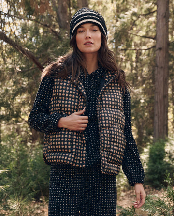 Woman standing outdoors in a forest, wearing a polka dot shirt, matching pants, a striped knit hat, and The Polar Vest. -- Black Paisley by THE GREAT. F26 TGO.