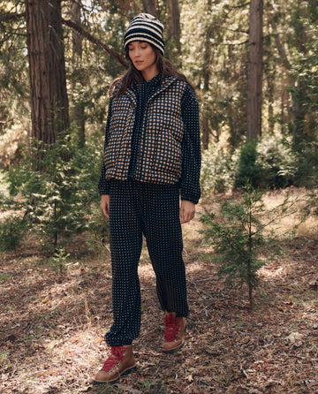 A person stands in a forest wearing The Polar Vest. -- Black Paisley by THE GREAT. F26 TGO over their jumpsuit, with a patterned beanie and brown hiking boots among tall, green trees.