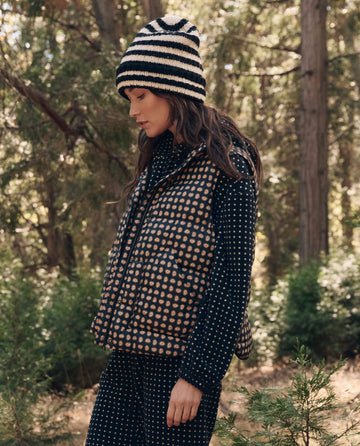 A person in a polka dot jacket and pants, wearing THE GREAT. F26 TGO The Polar Vest. -- Black Paisley and a striped knit hat, stands outdoors among tall trees and greenery.