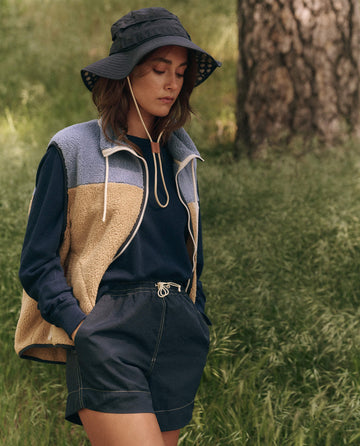 A person stylishly dressed in a plush bucket hat, THE GREAT. SP25 TGO's Creek Colorblock Terrain Vest, a long-sleeve shirt, and shorts stands outdoors near tall grass and a tree.