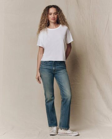 A person with curly hair stands against a beige backdrop, wearing THE GREAT. CORE KNITS The Crop Tee. Solid -- True White, blue jeans, and white sneakers.
