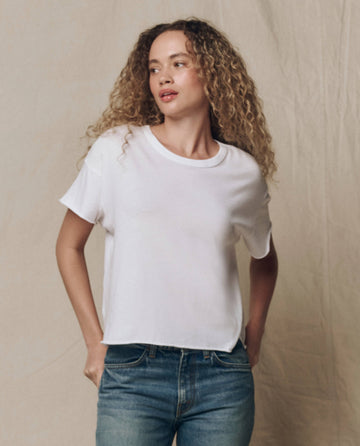A person with curly hair wears THE GREAT. CORE KNITS The Crop Tee. Solid -- True White and blue jeans, standing in front of a neutral, textured background.