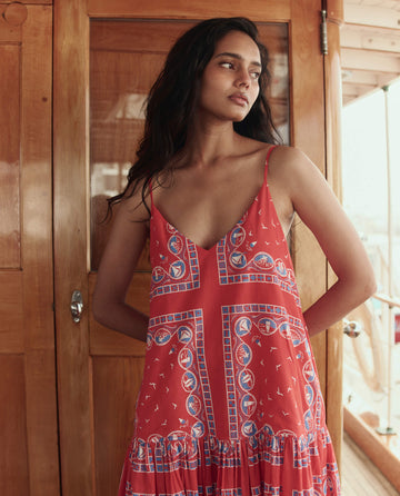 A woman stands indoors by a wooden door, wearing THE GREAT. SU26 The Drop Waist Dress -- Red Sailboat Bandana, a red sleeveless dress with blue and white geometric patterns.